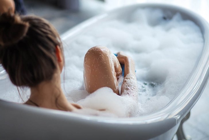 female massaging her legs with sponge in the tub. back view shot