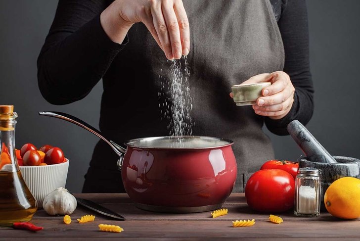 The chef preparations spaghetti and pasta, salt water, against a dark background, the concept of cooking. Woman salting water before cooking pasta fusilli